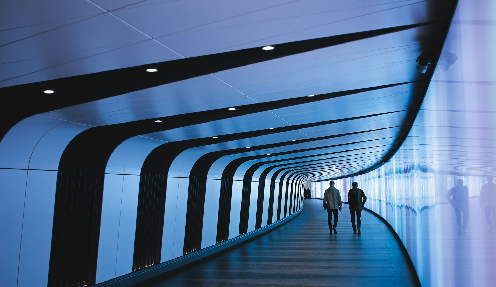 Two men walking down a long, curved hallway lit in blueish purple.